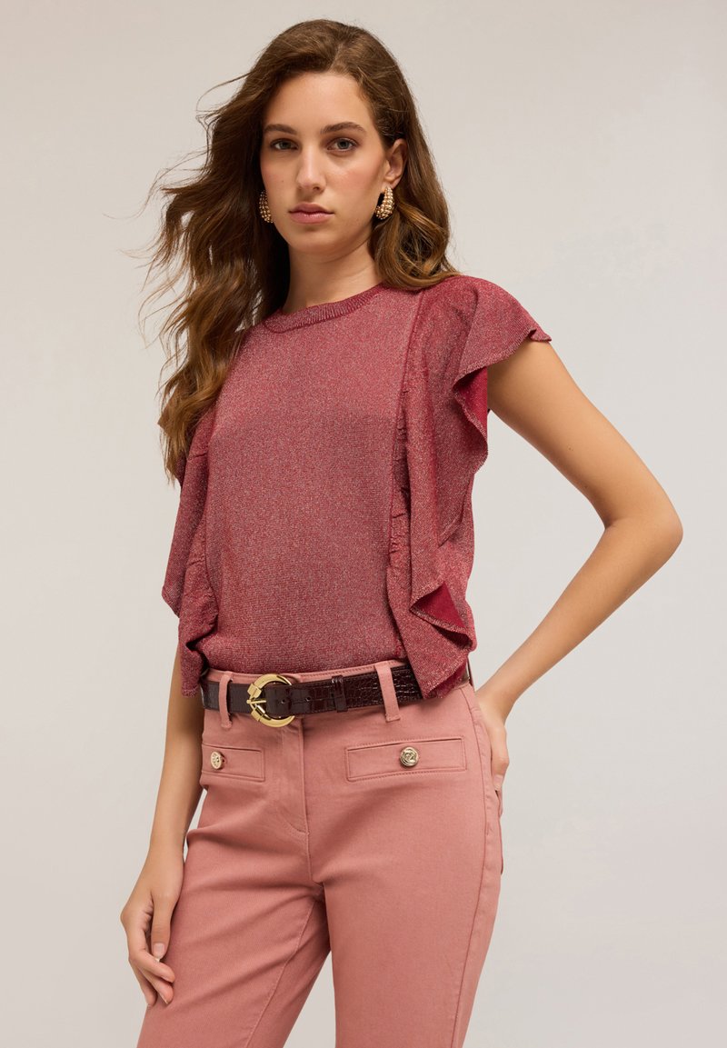 Una mujer con cabello ondulado lleva una blusa roja con volantes brillante, pantalones rosas con bolsillos con botones y un cinturón oscuro con hebilla dorada.