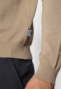 Μπεζ πλεκτό πουλόβερ με ριμπ μανσέτες και τελείωμα, με μικρή μαύρη ετικέτα λογότυπου στο πλάι. Φοριέται πάνω από σκούρο παντελόνι.