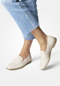 Beige leren loafers met een verhoogde naad, een platte hiel met een gouden accent en een brede band over de bovenkant. Gecombineerd met cropped denim jeans.