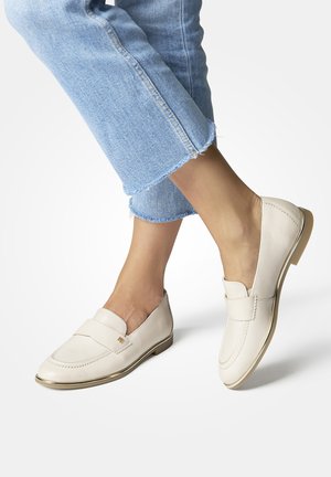 Beige leather loafers with a raised seam, a flat gold-accented heel, and a wide band across the upper. Worn with cropped denim jeans.