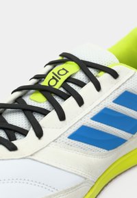 Bílá sportovní obuv s černými tkaničkami, zelenými akcenty a modrými pruhy. Má strukturovaný síťovaný svršek a žlutou gumovou podrážku.