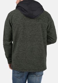 Sudadera de un verde oscuro texturizado con capucha negra, de corte relajado y mangas largas, combinada con unos jeans azules.