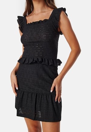 Kvinde iført en tætsiddende sort øjenlet kjole med flæser på skulderærmerne, firkantet halsudskæring, peplum-talje og flæset kant, står med hænderne i siden.