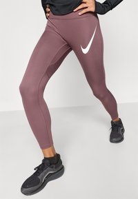 Κολάν με σχέδιο σύκου σε μωβ χρώμα, κατασκευασμένο από ελαστικό υλικό. Διαθέτει λευκό λογότυπο Nike στον μηρό και κομψό, εφαρμοστό σχέδιο. Εμφανίζονται μαύρα αθλητικά παπούτσια.