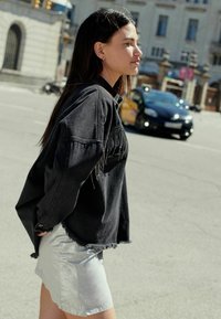 Juodas per didelis džinsinis švarkas su franelėmis, derinamas su šviesiai pilka sijonu. Fone - saulės apšviesta miesto aplinka.