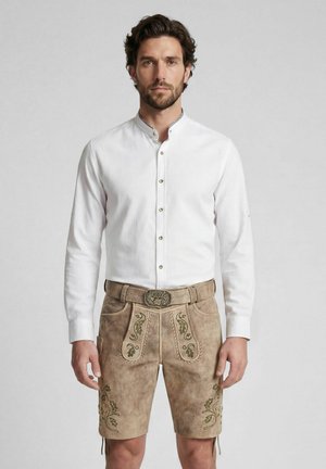 Mann trägt traditionelle beige bestickte Ledershorts mit Gürtel und weißem Hemd mit Knöpfen und steht vor schlichtem Hintergrund.