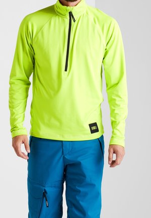 Homme portant un haut zippé vert néon à manches longues et un pantalon bleu avec fermeture éclair sur la poche, sur un fond clair uni.
