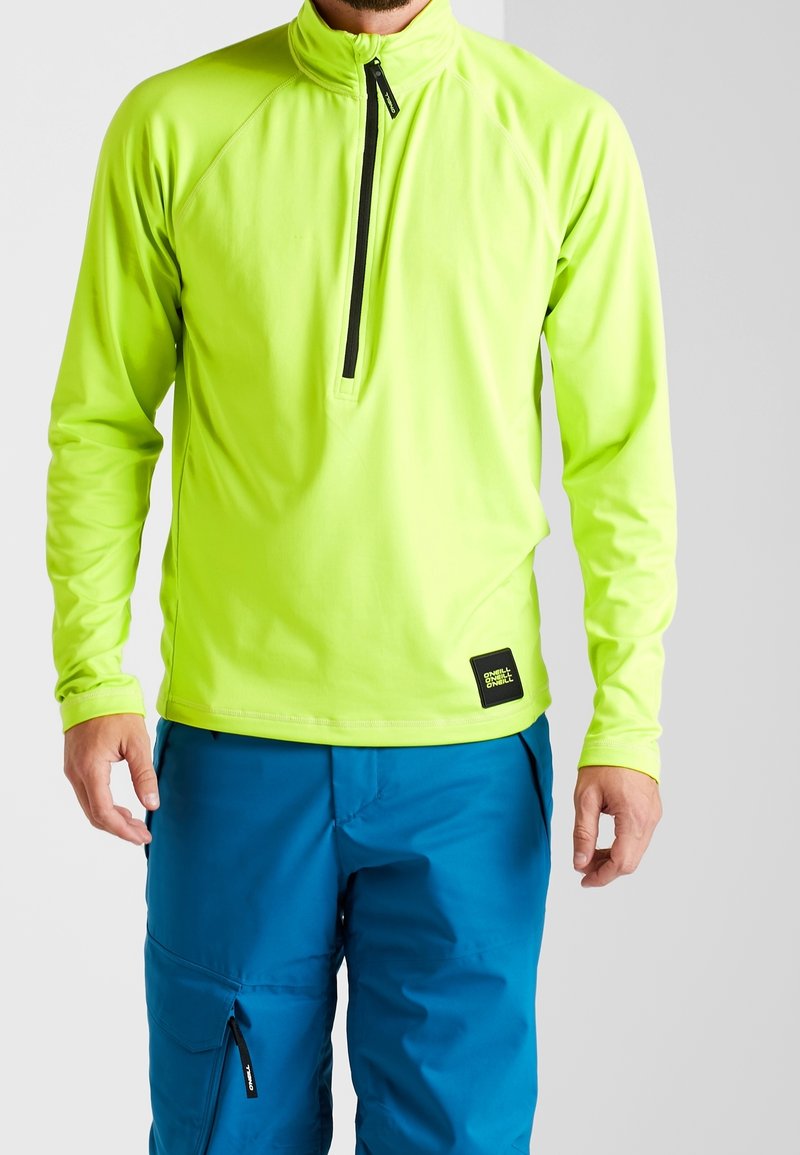 Homme portant un haut zippé vert néon à manches longues et un pantalon bleu avec fermeture éclair sur la poche, sur un fond clair uni.