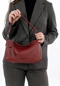 Rote Lederhandtasche mit geschwungenem Design, ausgestattet mit einem strukturierten Tragegurt, silbernen Hardware-Details und einem kleinen dekorativen Anhänger.