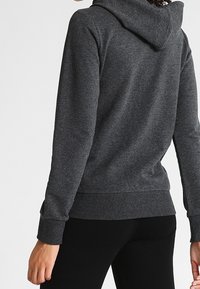 Sudadera gris con una tela texturizada, capucha ajustable con cordón, y puños acanalados. La vista trasera muestra un diseño entallado y un bolsillo canguro.