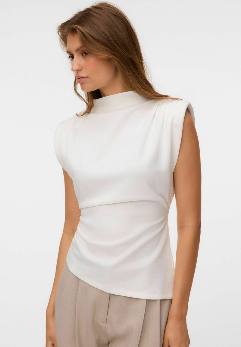 Jeune femme aux cheveux bruns portant un haut blanc sans manches avec des épaules rembourrées et un pantalon beige taille haute sur un fond uni.