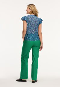 Zilā rakstaina blūze ar viļņainām piedurknēm, apvienota ar zaļām biksēm. Apģērbs izceļas ar košu paisley rakstu un vieglu audumu.