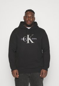 Black cotton hoodie featuring a front logo in silver with "Calvin Klein Jeans." It has a drawstring hood and ribbed cuffs and hem.
