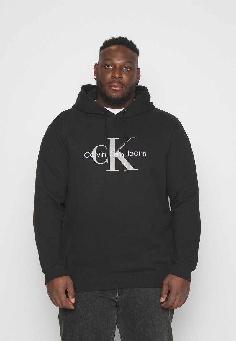 Black cotton hoodie featuring a front logo in silver with "Calvin Klein Jeans." It has a drawstring hood and ribbed cuffs and hem.