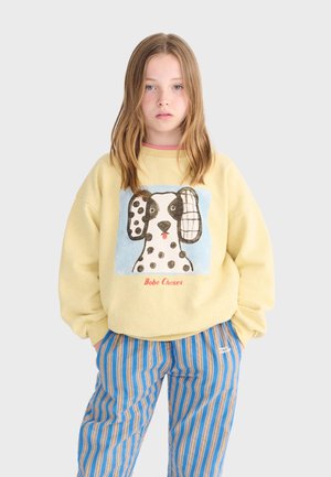 Nuori henkilö vaaleanruskein hiuksin, jolla on keltainen collegepaita, jossa on laikukas koira-aihe, ja siniruskearaidalliset housut.