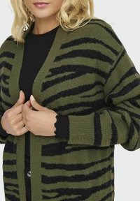 Zaļa un melna striped kardigana, izgatavota no mīksta adīta materiāla, ar brīvu piegriezumu un viļņu formas aproču detaļu. Atverama priekšējā daļa.