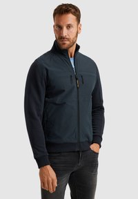 Giacca navy con tasche sul petto con zip, maniche testurizzate e polsini a coste. Presenta un collo alto e un design slanciato.