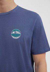 Man met een blauwe t-shirt met een ronde patch waarop staat "Somewhere in the Urban Universe" op de linkerkant van de borst.