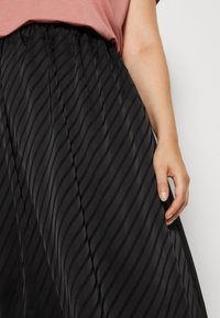 Svart plissé skjørt med diagonale striper, laget av glatt stoff. Livstykket er elastisk, og teksturen reflekterer lyset subtilt.