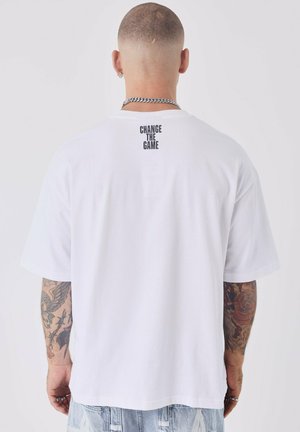 Camiseta de algodón blanca con mangas cortas, que presenta el texto "CAMBIA EL JUEGO" impreso en negro en la parte superior de la espalda. Textura suave.
