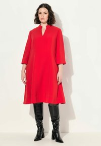 Vestido rojo a la altura de la rodilla con escote en forma de llave y mangas de tres cuartos. Combinado con botas negras de caña alta y tacón alto. Textura suave.