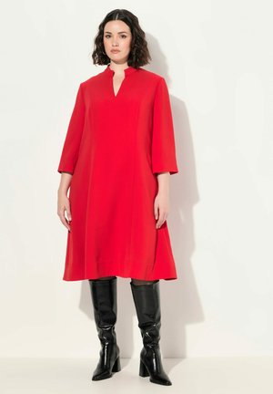 Vestido rojo a la altura de la rodilla con escote en forma de llave y mangas de tres cuartos. Combinado con botas negras de caña alta y tacón alto. Textura suave.