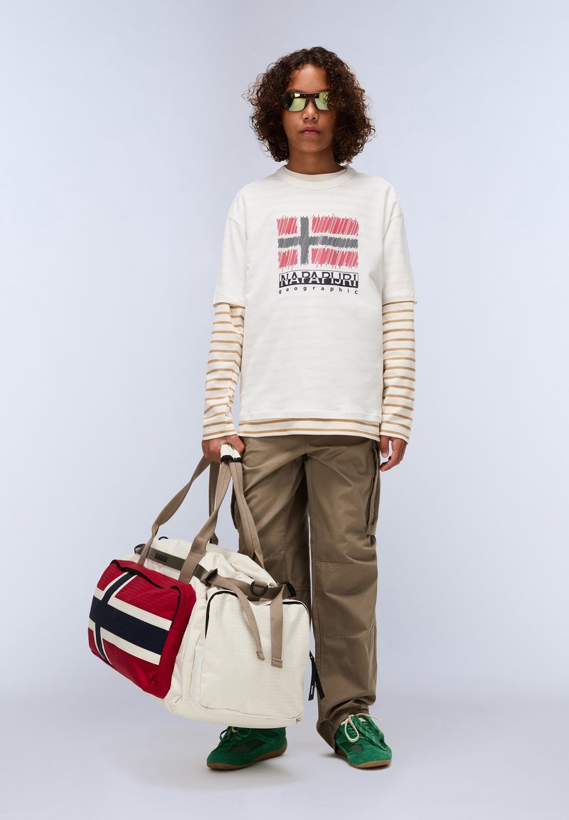 Model wears a cream graphic T-shirt with Norwegian flag design, beige striped long-sleeve underneath, khaki pants, and green shoes. Carrying a large duffel bag with red and navy accents.