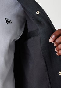 Svart jakkeinnside med en jevn, matt tekstur; har et hvitt indre fôr og en liten brodert logo. Detaljer med knapp og elastisk bånd synlige.