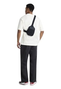 Black sling bag with zipper pockets, worn over a light-colored t-shirt. The bag features a smooth texture and adjustable strap.