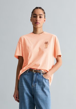 Jeune femme aux cheveux tressés portant un t-shirt pêche avec un petit motif, rentré dans un jean taille haute bleu avec une ceinture marron.