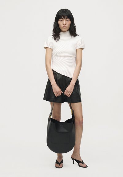 Un mannequin se tient devant un fond uni, vêtu d’un haut blanc côtelé, d’une jupe noire et de tongs à talons, et tient un grand sac noir porté à l’épaule.