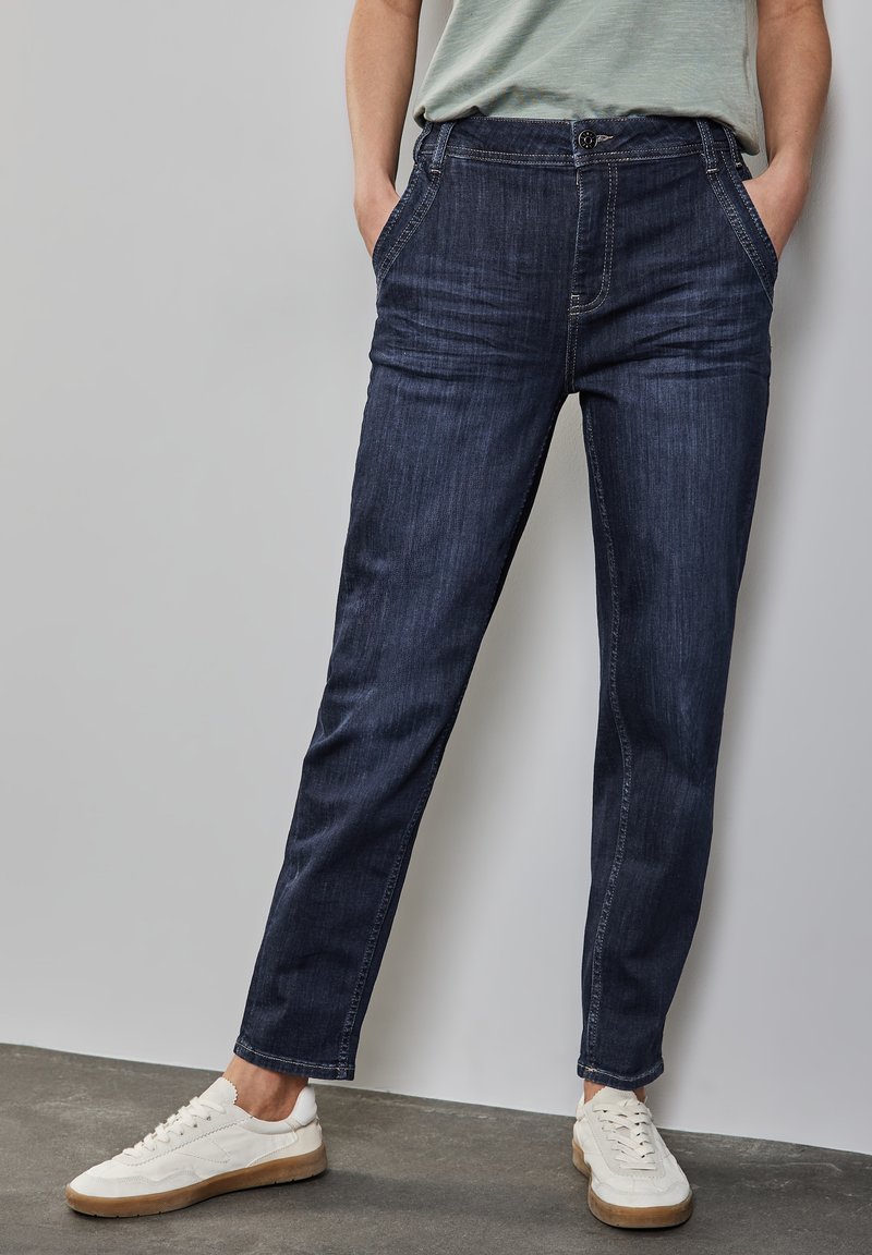 Jean en denim bleu foncé avec une coupe droite, comprenant des passants de ceinture, des poches latérales et une texture subtile, associés à des baskets blanches.