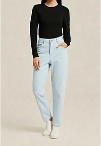 Kobieta ubrana w czarną bluzkę z długim rękawem, jasnoniebieskie dżinsy z wysokim stanem oraz białe trampki na jednolitym tle.