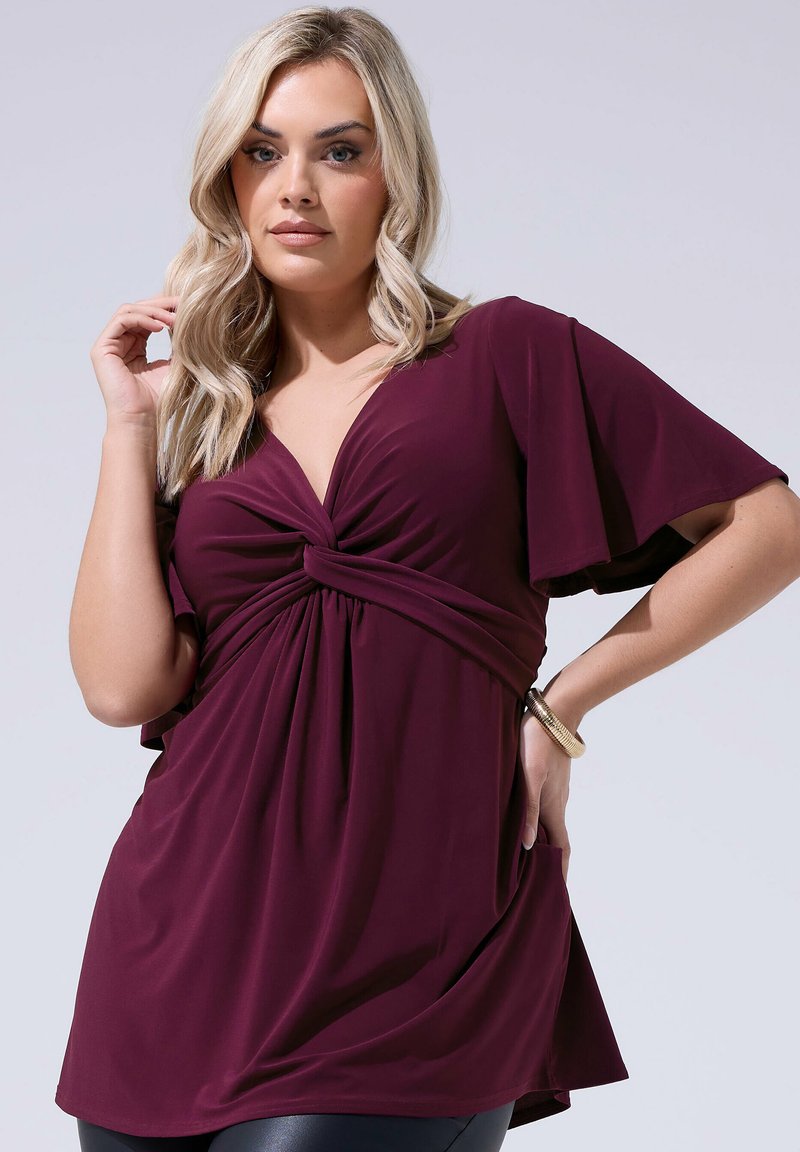 Mujer rubia que lleva un vestido color púrpura oscuro con nudo delantero, mangas de vuelo y una pulsera dorada, posando contra un fondo claro.