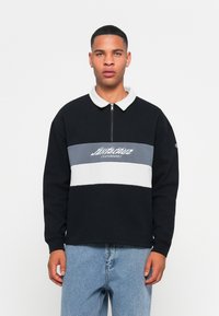 Jonge man in een zwarte pullover met wit en grijs horizontale strepen en een ritskraag, gecombineerd met lichtblauwe jeans, die recht vooruit kijkt.