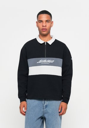 Jonge man in een zwarte pullover met wit en grijs horizontale strepen en een ritskraag, gecombineerd met lichtblauwe jeans, die recht vooruit kijkt.
