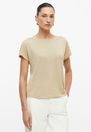 Femme aux cheveux foncés portant un haut beige à manches courtes et un pantalon blanc, debout devant un fond blanc uni.