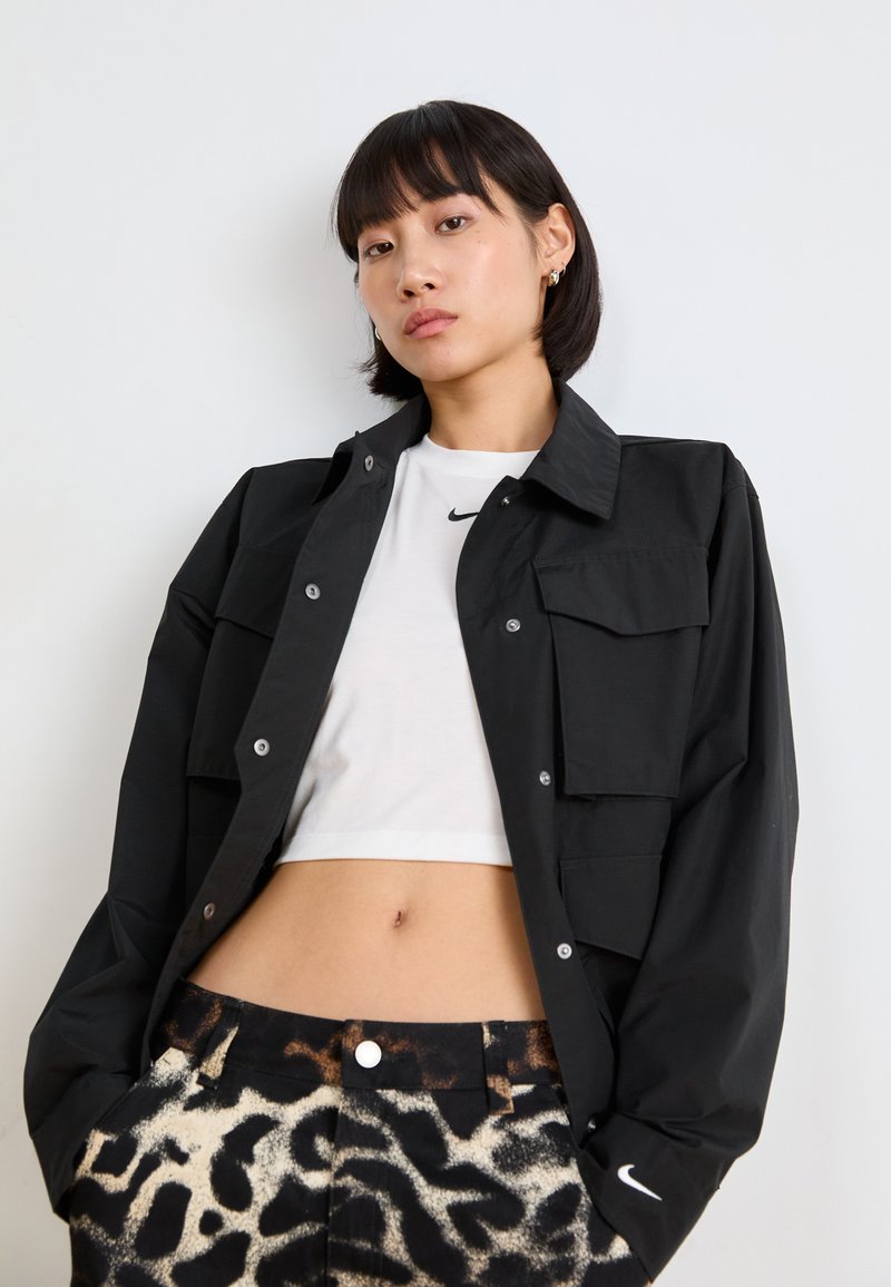Veste noire à boutons, avec des poches à rabat, portée sur un top court blanc. Associée à un pantalon à motifs noir et beige avec une fermeture à bouton.