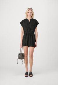 Robe courte noire à manches courtes avec deux poches poitrine, taille élastique et coupe décontractée. Le modèle tient un petit sac à motifs et porte des sandales noires.