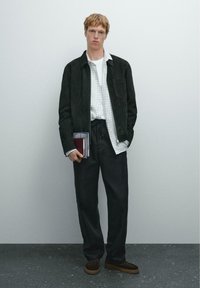 Schwarze Wildlederjacke über einem weiß karierten Hemd, kombiniert mit dunklen Hosen und braunen Slippern. Hält ein Notizbuch und einen Reisepass.