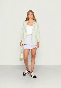 Blazer verde-claro sobre um top cropped branco, emparelhado com uma saia lilás e sandálias pretas. Os acessórios incluem uma pequena bolsa verde.