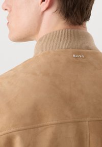 Smeđi nubuk jakna s rebrastim ovratnikom. Srebrni "BOSS" logotip smješten blizu vrata. Glatka tekstura s čistim krojem.