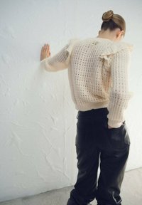 Suéter de punto color crema con detalles de volantes, combinado con pantalones de cuero negro. El fondo presenta una pared blanca con textura.