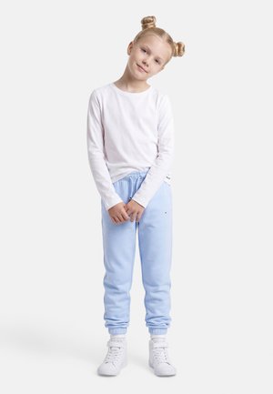 Ung pige med blondt hår i knolde iført en hvid langærmet trøje, lyseblå joggingbukser og hvide sneakers, stående mod en hvid baggrund.