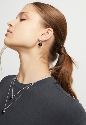Boucles d'oreilles en perles noires et colliers en argent superposés portés avec une chemise sombre. Cheveux attachés, mettant en valeur les accessoires et le décolleté.