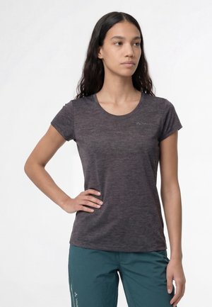 Frau mit langen dunklen Haaren, die ein eng anliegendes dunkelgraues Kurzarmshirt und türkisfarbene Hose trägt und mit einer Hand in der Hüfte steht.