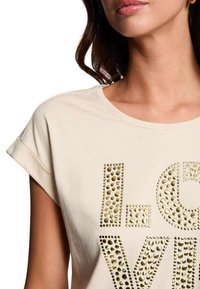 Lichtbeige t-shirt met opgerolde mouwen, voorzien van gouden gestifte letters. Zachte katoen, ronde halsontwerp, gladde structuur, casual pasvorm.