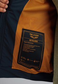 Marineblauer Softshell-Jacke mit orangenem Netzfutter. Zu den Merkmalen gehören wasserdichtes Material, gerippte Bündchen, Handtaschen und eine innere mit Reißverschluss versehenen Tasche.