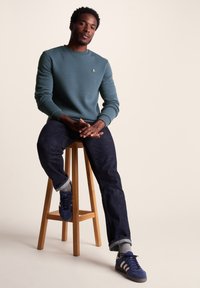 Teal sweatshirt with ribbed cuffs, wooden stool, dark denim jeans, grey socks, and blue sneakers with white stripes. Minimalistic logo detail.