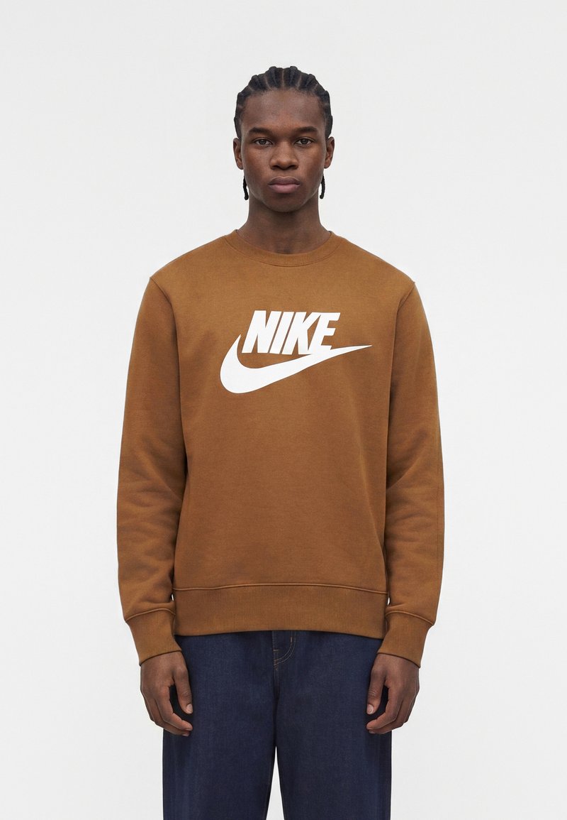 Junger Mann mit geflochtenem Haar, der einen braunen Nike-Sweatshirt und dunkelblaue Jeans trägt, steht vor einem weißen Hintergrund.